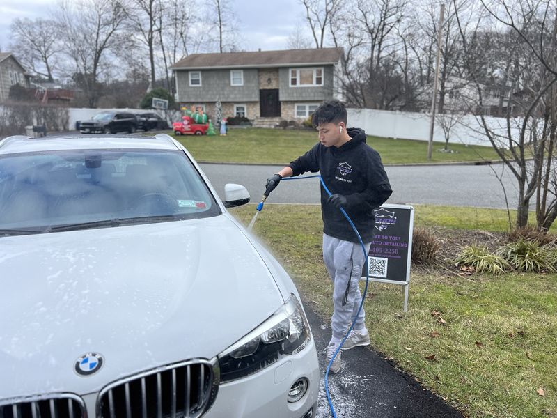 Mobile hand car wash in Charlotte, NC: Detailer rinsing a white BMW X3 in a driveway with a Motion Mobile Detailing sign behind him