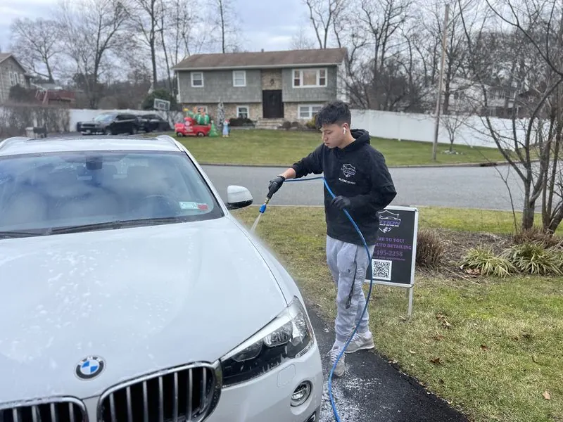 Premium mobile car detailing in Greensboro, NC: Detailer rinsing a white BMW X3 in a driveway with a Motion Mobile Detailing sign behind him