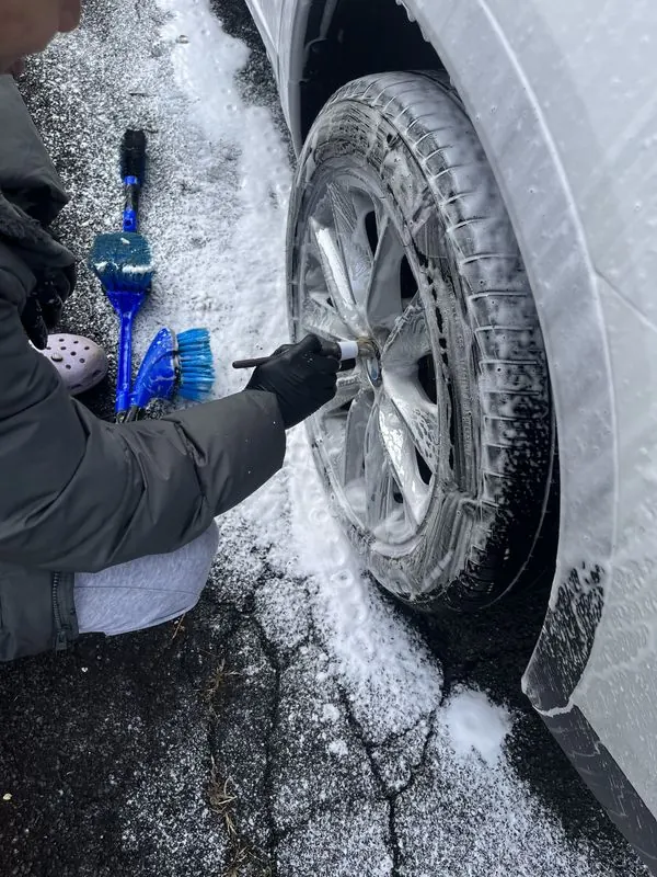 Detailer scrubbing a wheel with a soft brush through thick foam during a mobile hand wash on a cold day